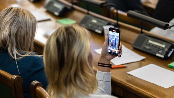 Депутат Сейма Латвии фотографирует на смартфон выступление премьер-министра Латвии Кришьяниса Кариньша - Sputnik Латвия